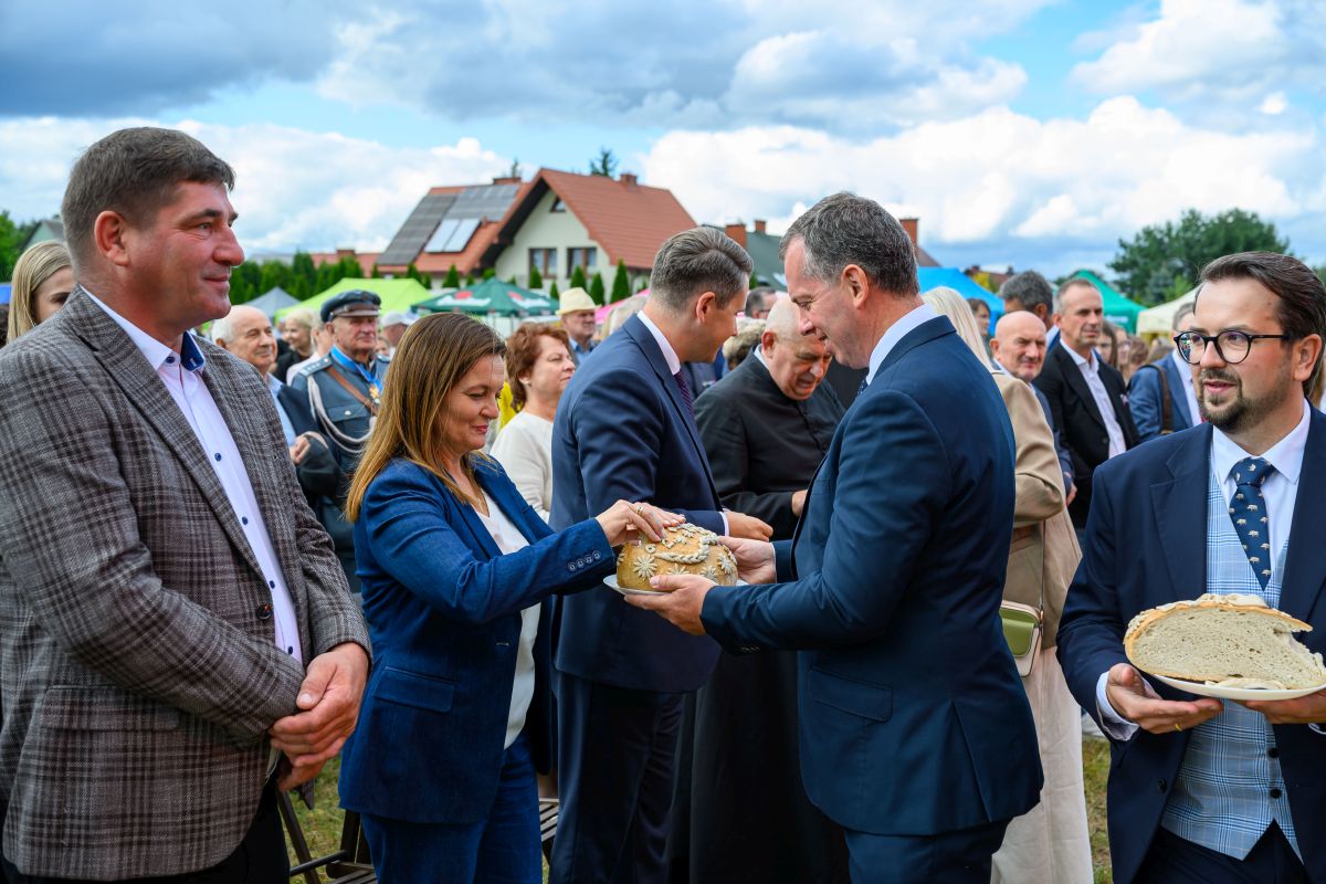 Na zdjęciu grupa ludzi w eleganckich ubraniach dzielących chleb. Tło to niebo z chmurami i domy z czerwonymi dachami. Wydarzenie odbywa się na świeżym powietrzu.