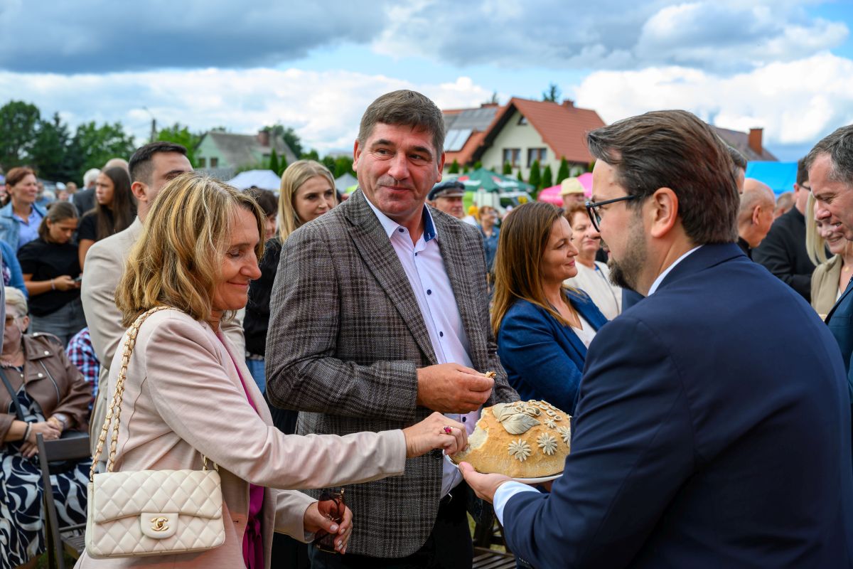 Grupa osób na zewnątrz uczestniczy w wydarzeniu. Kobieta w jasnym płaszczu łamie chleb. W tle domy i pochmurne niebo.