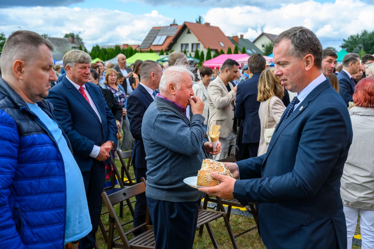 Grupa ludzi na zewnątrz, jeden mężczyzna oferuje chleb. W tle domy i kolorowe namioty. Uczestnicy ubrani elegancko.
