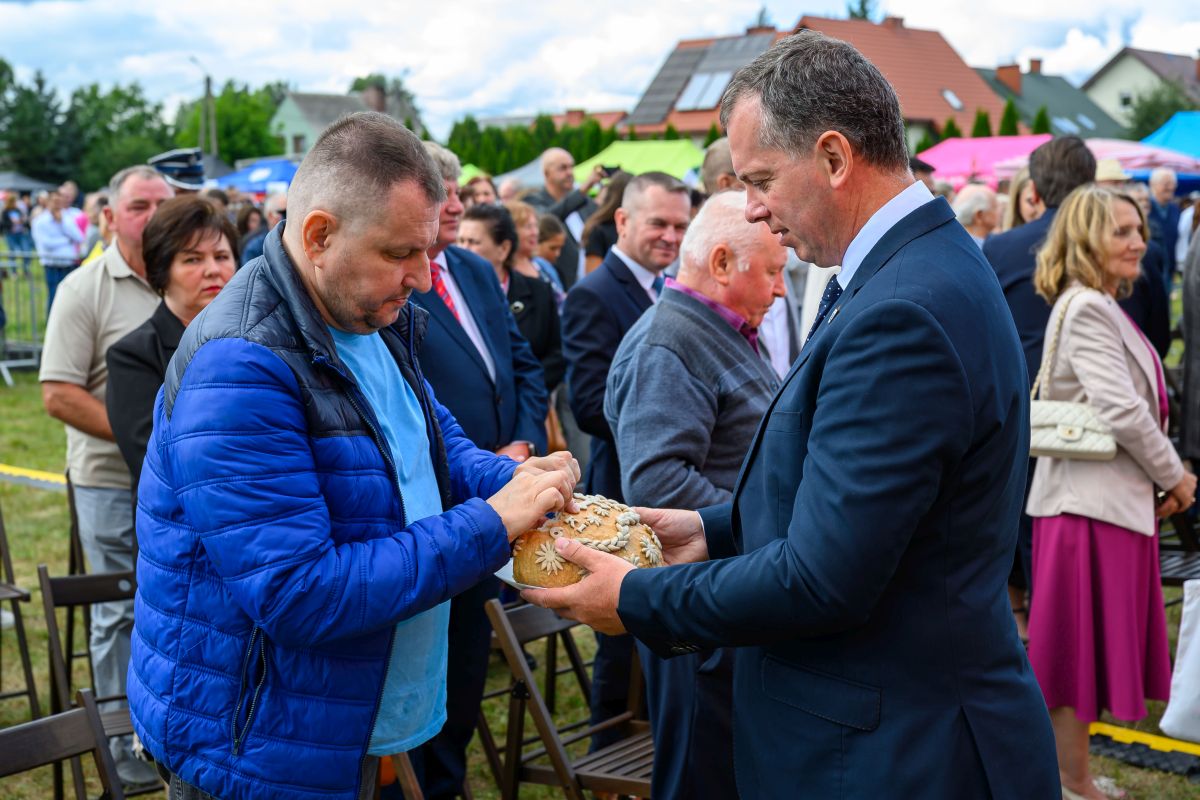 Mężczyźni ubrani w garnitur i niebieską kurtkę dzielą się chlebem na zewnątrz. Tło to tłum ludzi i kolorowe parasole. Atmosfera jest uroczysta.