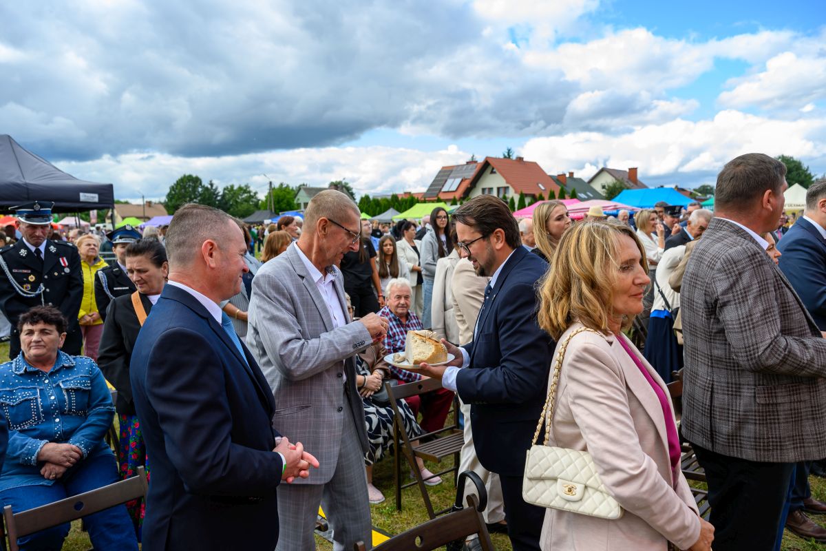 Grupa ludzi w eleganckich strojach uczestniczy w wydarzeniu plenerowym. Niebo jest pochmurne, a w tle widać namioty i domy. Osoba w garniturze wręcza kawałek chleba innemu uczestnikowi.
