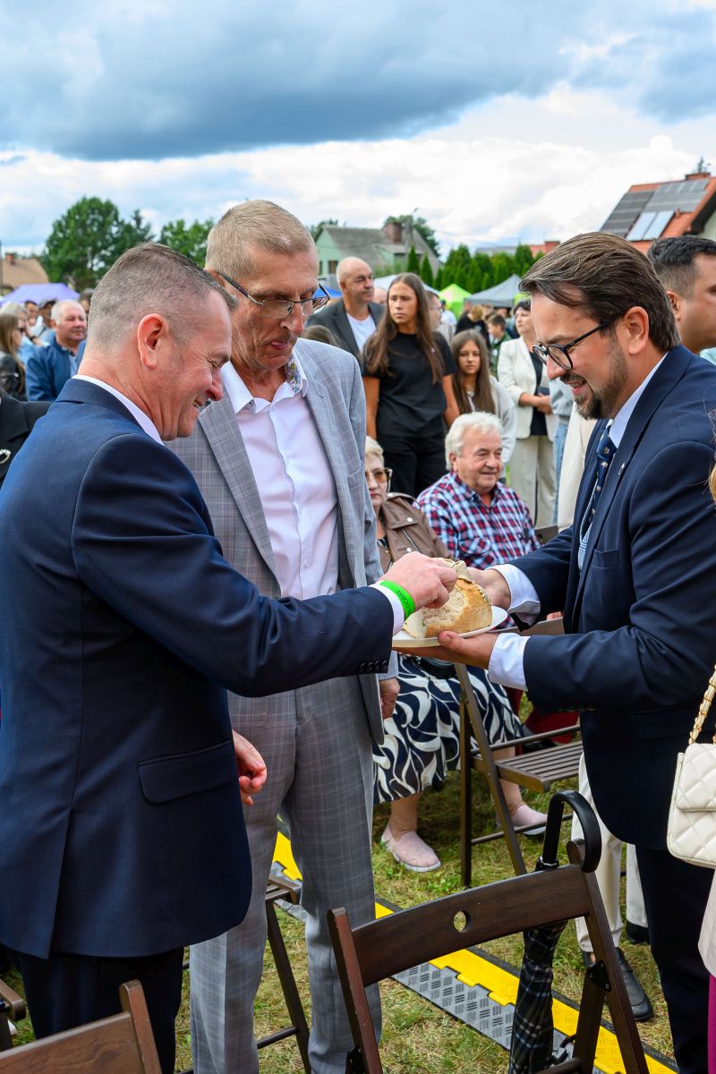 Trzej mężczyźni w eleganckich garniturach dzielą się chlebem. W tle tłum na zewnątrz, niebo pokryte chmurami. Atmosfera wydarzenia formalnego.