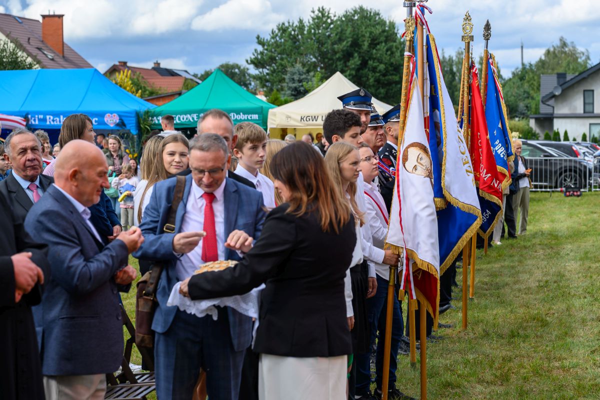 Grupa ludzi na uroczystości plenerowej, w tle kolorowe namioty i flagi. Osoby są elegancko ubrane. Tło to zielona trawa i drzewa.