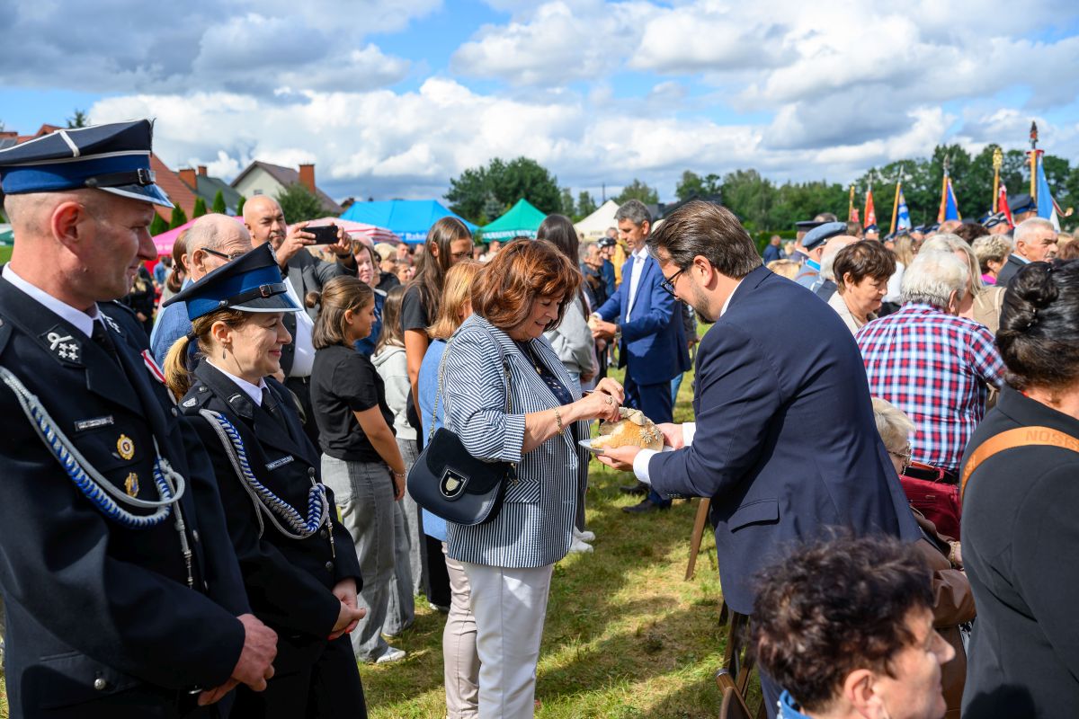 Na zdjęciu tłum ludzi na zewnątrz, w tym osoby w mundurach i kobieta krojąca chleb. W tle niebieskie niebo i namioty. Wydarzenie odbywa się na trawie.