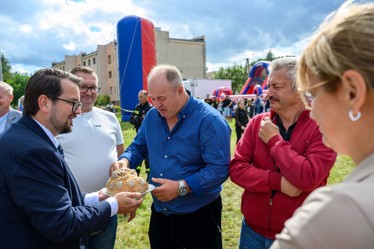 Grupa mężczyzn na trawie, jeden trzyma ozdobny bochenek chleba. W tle niebiesko-czerwona dmuchana atrakcja. Budynek pod chmurami.