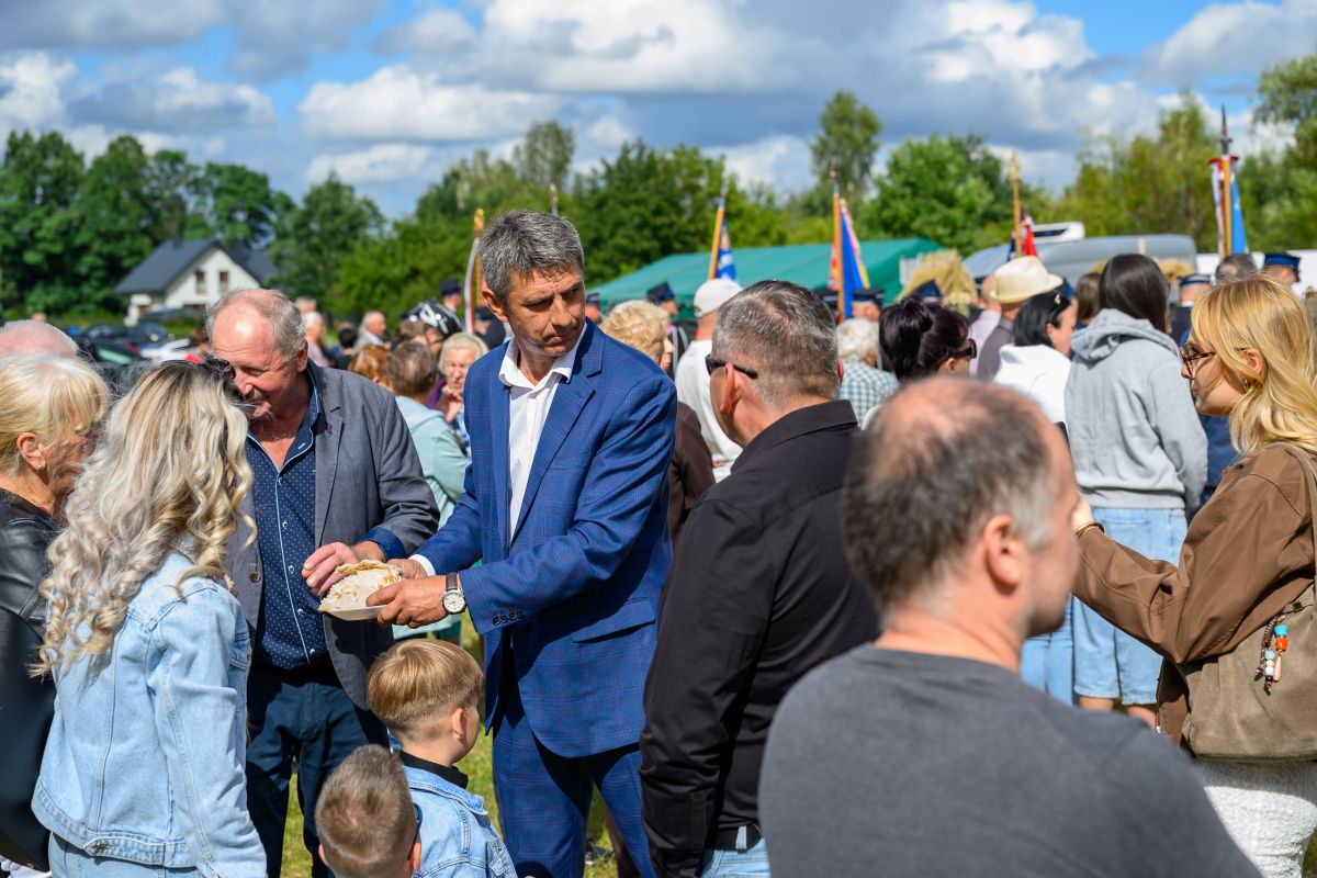 Na zdjęciu jest grupa osób na zewnątrz podczas wydarzenia. Mężczyzna w granatowym garniturze trzyma chleb. Tło jest zielone z flagami i niebieskim niebem.