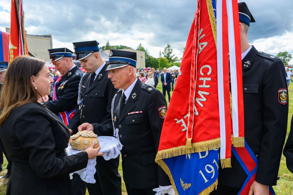 Ceremonia strażacka na świeżym powietrzu, mężczyźni w mundurach obok kobiety z chlebem. Czerwone sztandary dominują scenę. Tło to chmury i zielone drzewa.