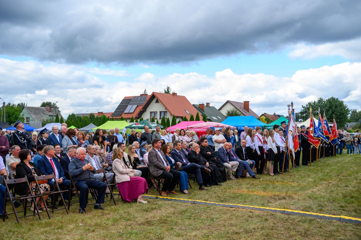 Na zdjęciu widać ludzi siedzących na krzesłach na zewnątrz. W tle są kolorowe namioty i domy. Niebo jest pochmurne.