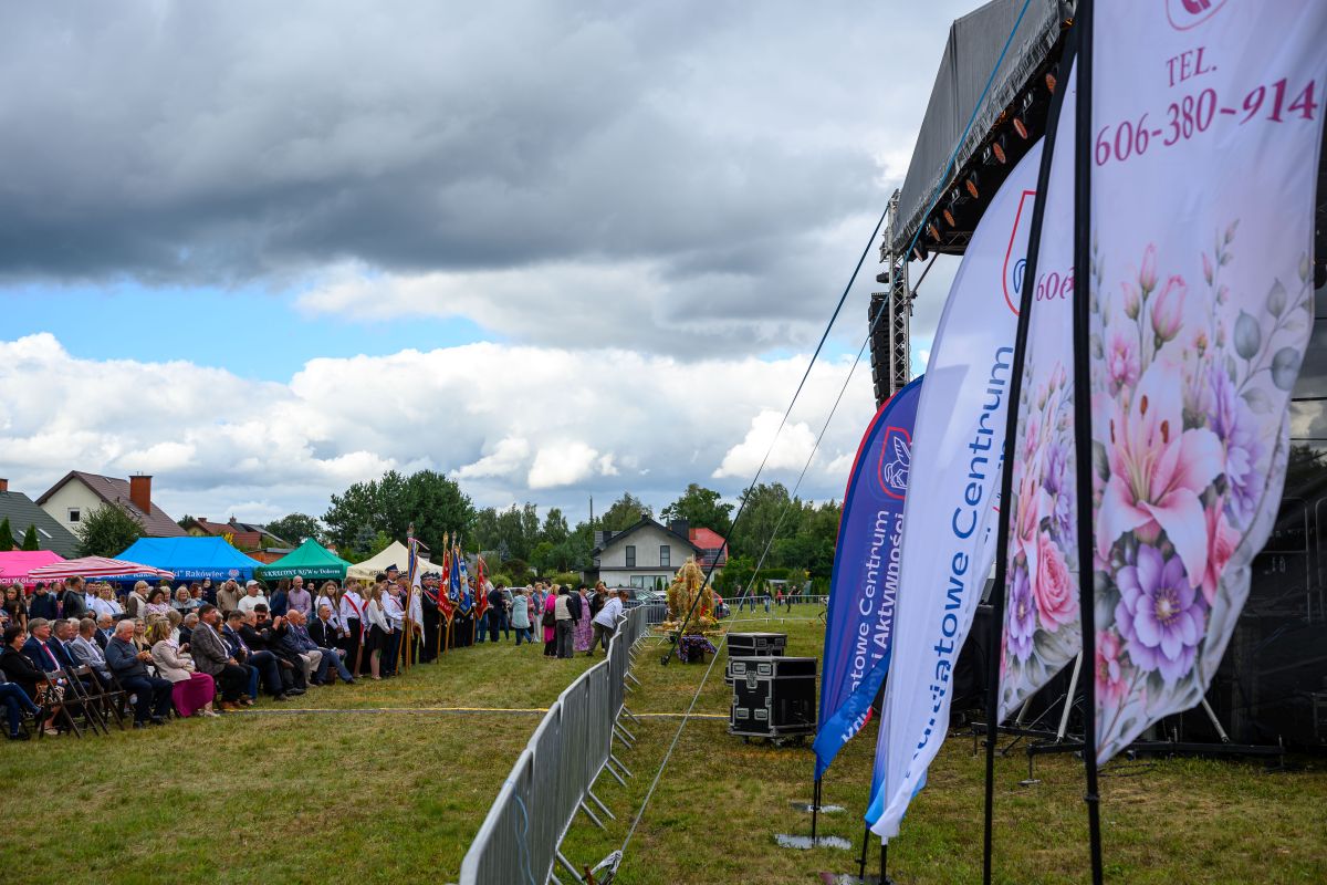 Plenerowe wydarzenie z tłumem ludzi, kolorowymi namiotami i flagami na zielonej trawie. W tle domy i pochmurne niebo. Scena po prawej stronie.