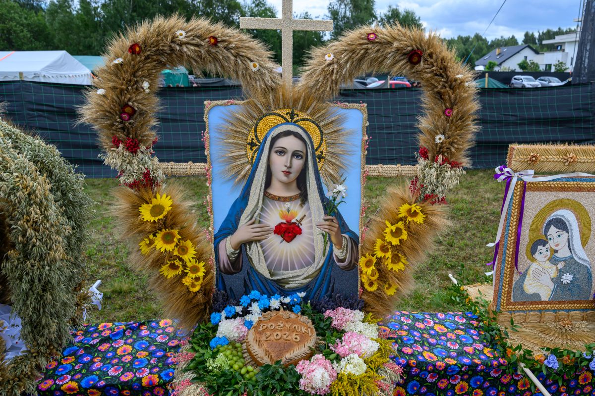Obraz przedstawia wizerunek Maryi z otoczką z kłosów i słoneczników. Na dole kompozycja z kolorowych kwiatów i napisem. W tle zielone drzewa i namioty.