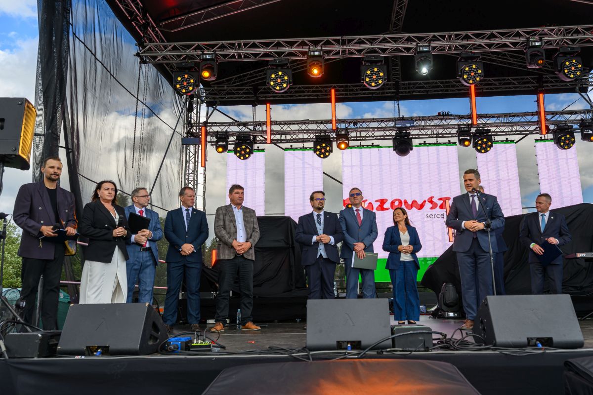 Grupa ludzi stoi na scenie pod czarnym zadaszeniem. Tło to wielki ekran z napisem w czerwieni. Prowadzący trzyma mikrofon.