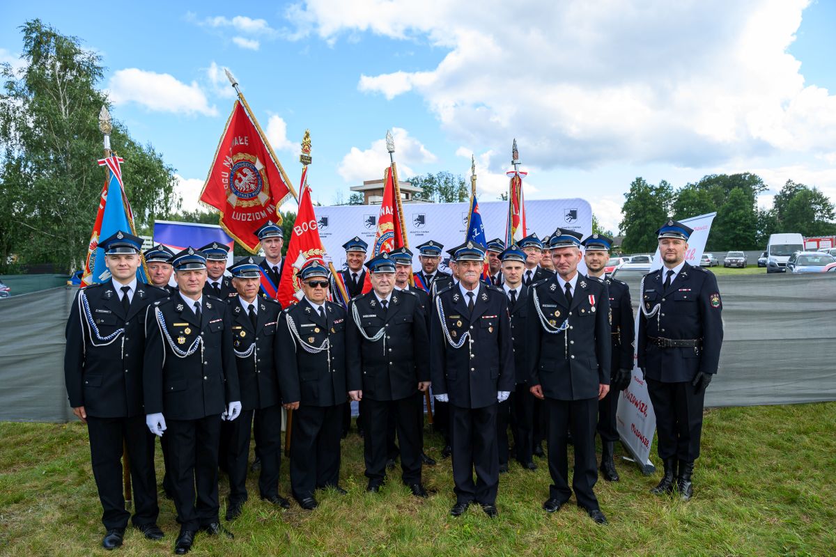 Grupa strażaków w czarnych mundurach z niebieskimi czapkami stoi na trawie. Za nimi widać kilka kolorowych flag. W tle błękitne niebo i drzewa.