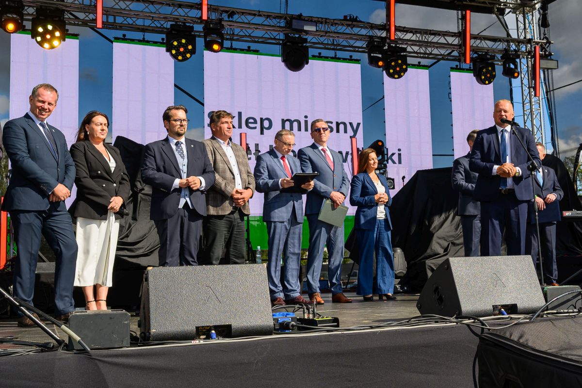 Na scenie stoi grupa osób w eleganckich strojach. W tle jasne ekrany z napisami. Na scenie sprzęt nagłośnieniowy.