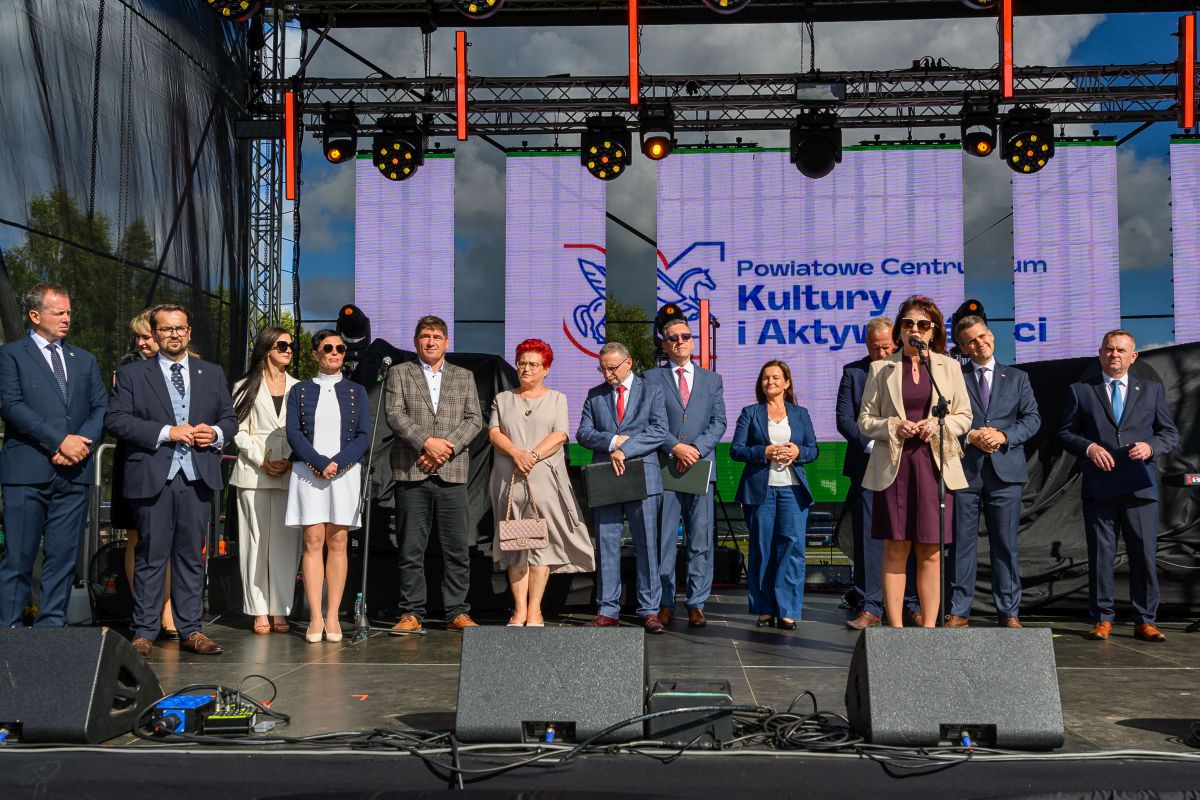 Na scenie stoi grupa osób w eleganckich strojach. Tło to duży ekran z napisem Powiatowe Centrum Kultury. Scena oświetlona jest jasnym światłem dziennym.