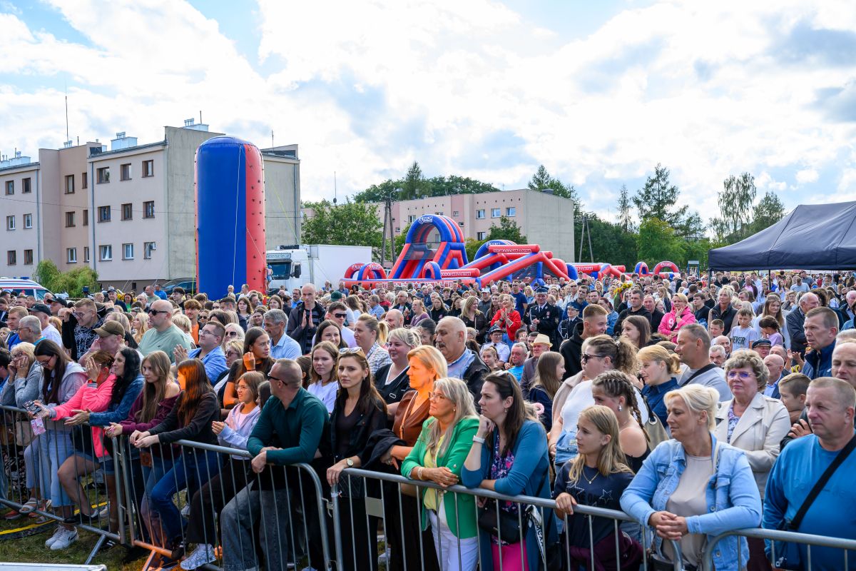 Tłum ludzi na zielonej łące przy kolorowych dmuchańcach. W tle bloki mieszkalne i drzewa. Niebieskie niebo z białymi chmurami.