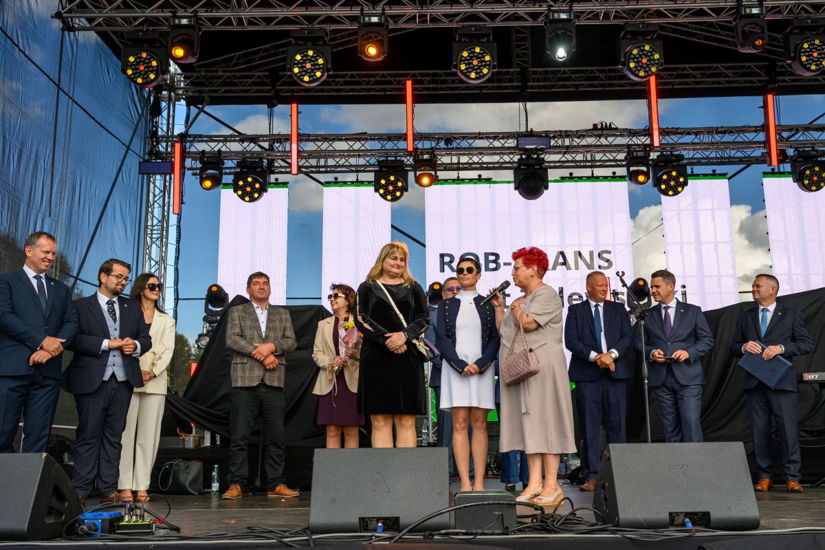 Na scenie stoi grupa ludzi w eleganckich ubraniach. Tło stanowi duży ekran z napisem i światła sceniczne. Niebo jest częściowo widoczne.