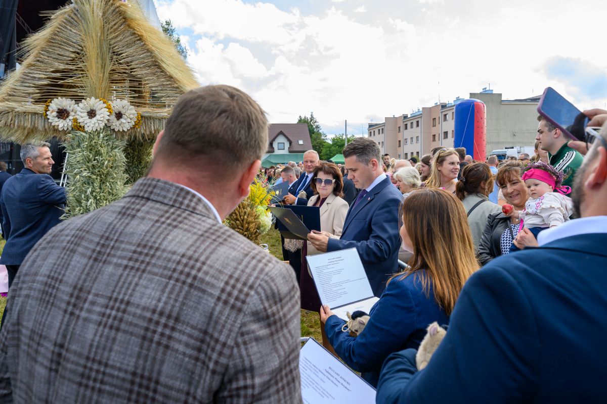 Na zdjęciu ludzie zgromadzeni wokół ozdoby ze zbóż. W tle budynki, niebo z chmurami, dominują kolory niebieski i zielony. Atmosfera wydarzenia plenerowego.