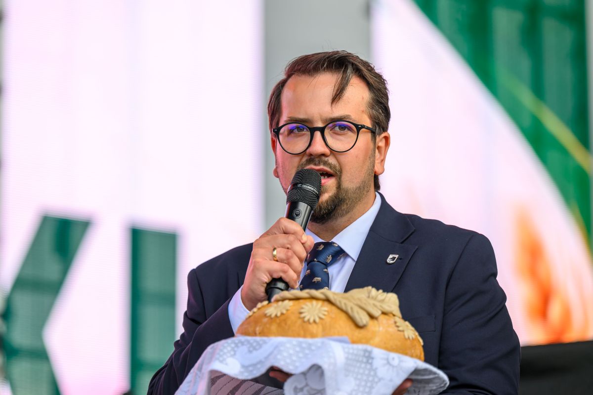 Mężczyzna w okularach trzyma chleb na scenie, mówi do mikrofonu. Ubrany w garnitur, w tle zielono-białe wzory. Na chlebie dekoracje z ciasta.