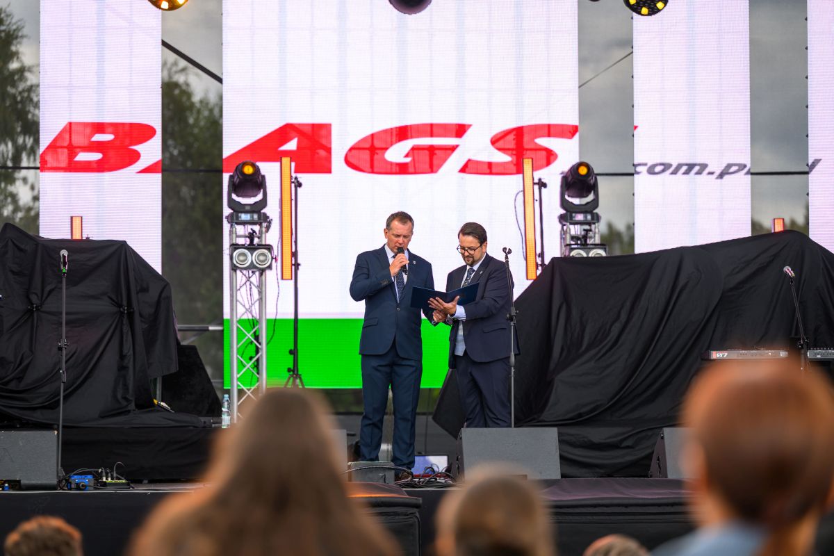 Dwóch mężczyzn w garniturach na scenie z mikrofonem i notatnikiem. Tło z jasnymi ekranami. Widownia przed sceną.