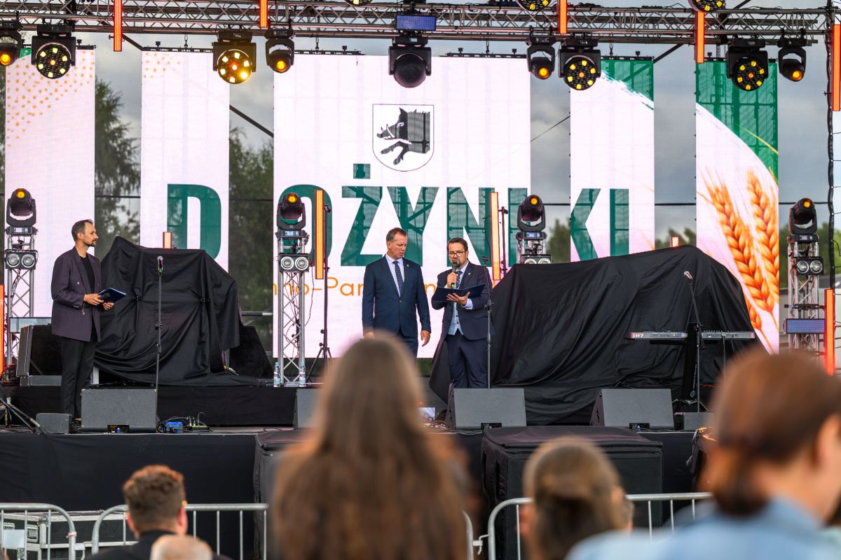 Na scenie, na tle dużego ekranu LED, dwaj mężczyźni w garniturach przemawiają. Ekran pokazuje napis Dożynki i symbol kłosa. Scena jest oświetlona reflektorami.