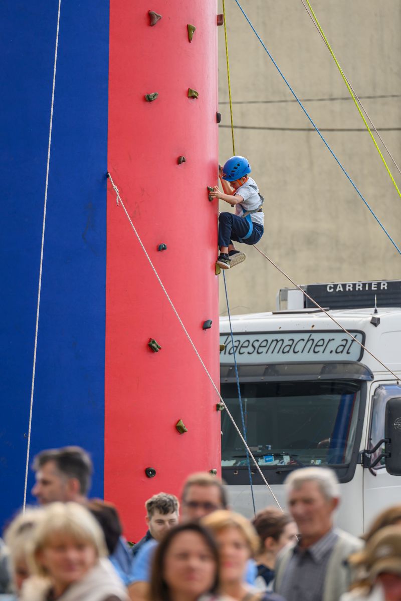 Chłopiec wspina się na ściankę wspinaczkową w kolorach czerwonym i niebieskim. Ma na sobie niebieski kask. W tle stoi biały samochód ciężarowy.