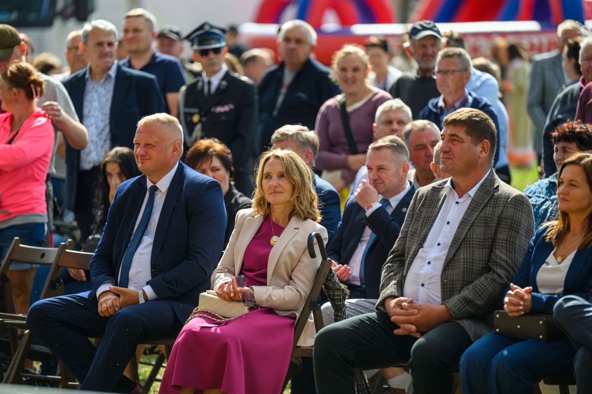 Grupa osób siedzi na składanych krzesłach na zewnątrz. Centralna kobieta w różowej sukience, mężczyzna obok w niebieskim garniturze. Tło z rozmytą publicznością i elementami w kolorze czerwonym i niebieskim.