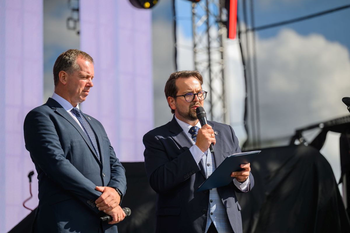 Dwóch mężczyzn w garniturach na scenie; jeden przemawia do mikrofonu, trzymając notatnik. Tło to jasne niebo i konstrukcja sceniczna. Słoneczne oświetlenie podkreśla formalność wydarzenia.
