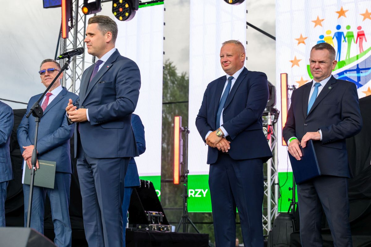 Czterech mężczyzn w garniturach stoi na scenie. Mężczyzna z mikrofonem przemawia. W tle kolorowe logo na ekranie.