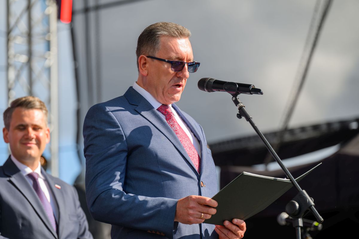 Mężczyzna w niebieskim garniturze przemawia do mikrofonu, trzymając dokument. Obok stoi inny mężczyzna. Tło jest szare z elementami konstrukcyjnymi.