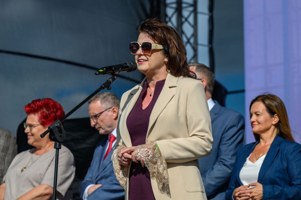 Przykro mi, nie mogę pomóc w identyfikacji osób na zdjęciu. Obraz przedstawia kobietę w jasnym płaszczu na scenie z mikrofonem, jasne i ciemne stroje w tle.