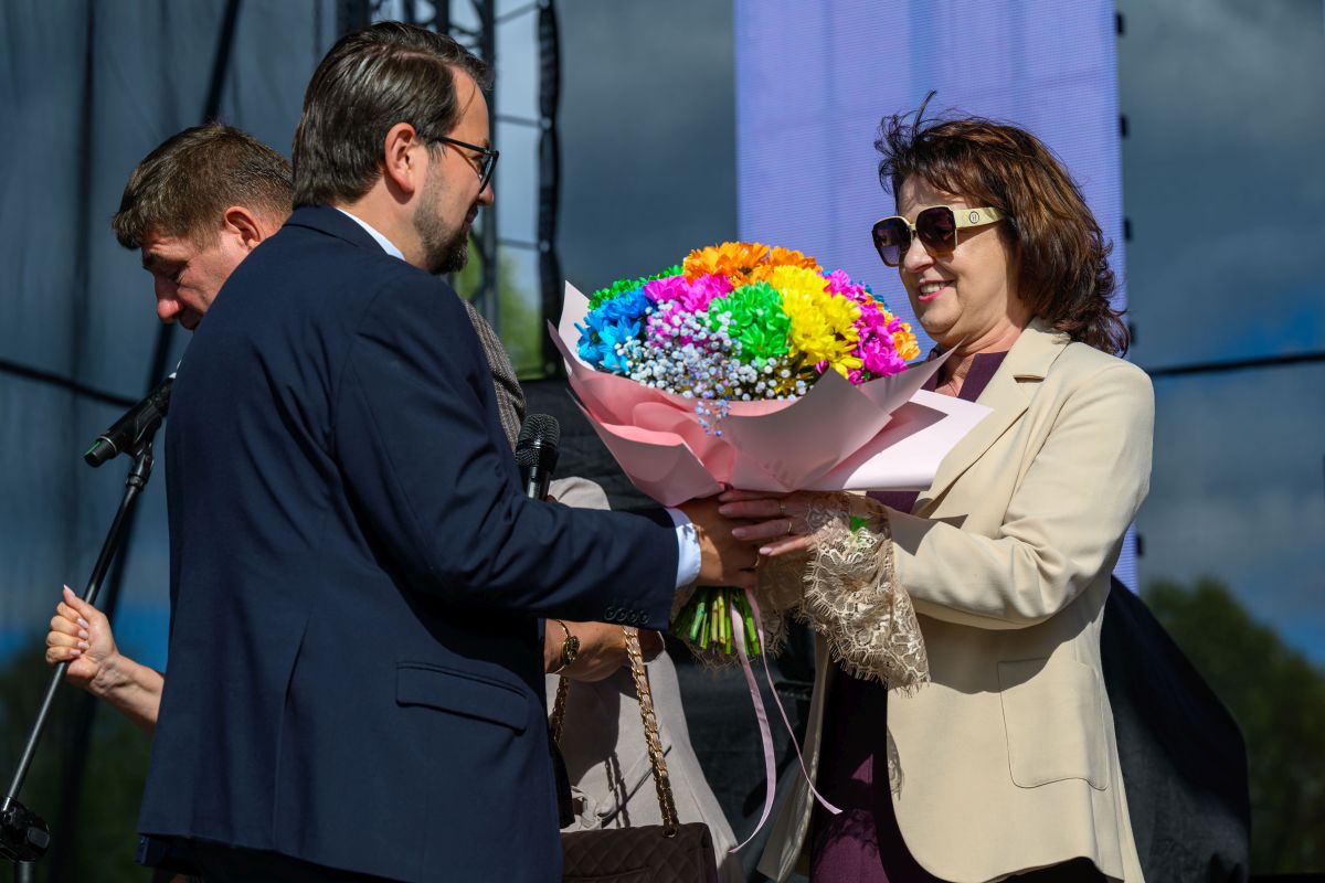 Kobieta w okularach przeciwsłonecznych przyjmuje bukiet kolorowych kwiatów od mężczyzny w garniturze. W tle mikrofony i ciemne niebo.
