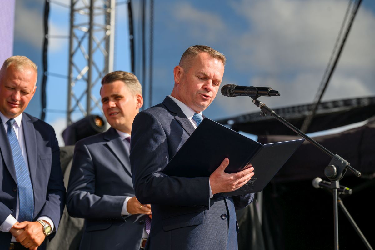 Trzech mężczyzn w garniturach, jeden z mikrofonem i dokumentem, tło z jasnym niebem i konstrukcją sceny.