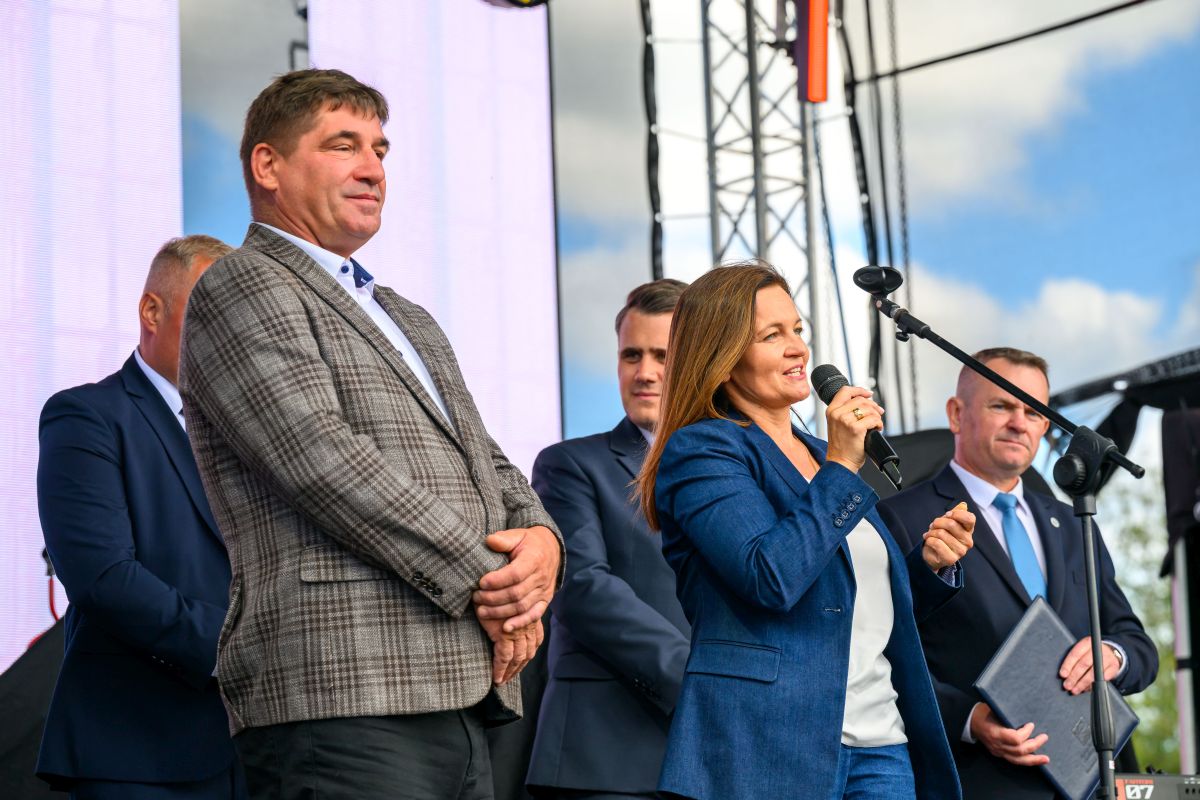 Grupa ludzi na scenie; kobieta w niebieskiej marynarce mówi do mikrofonu. Jasne niebo w tle, widoczna struktura nośna.