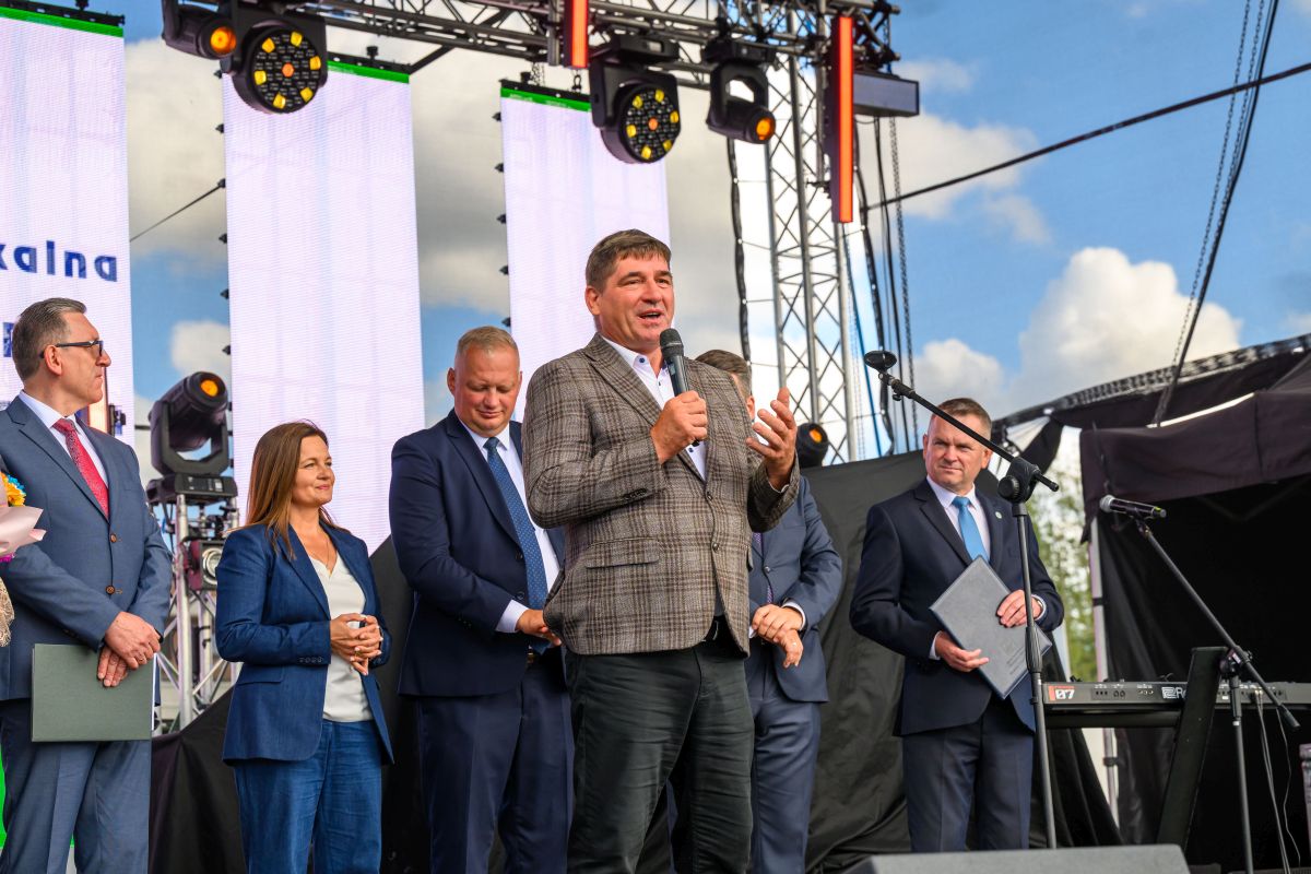 Na scenie stoi mężczyzna w marynarce, przemawiający do mikrofonu. W tle są jasne ekrany LED i konstrukcja sceniczna. Obok niego stoi grupa ludzi w garniturach.
