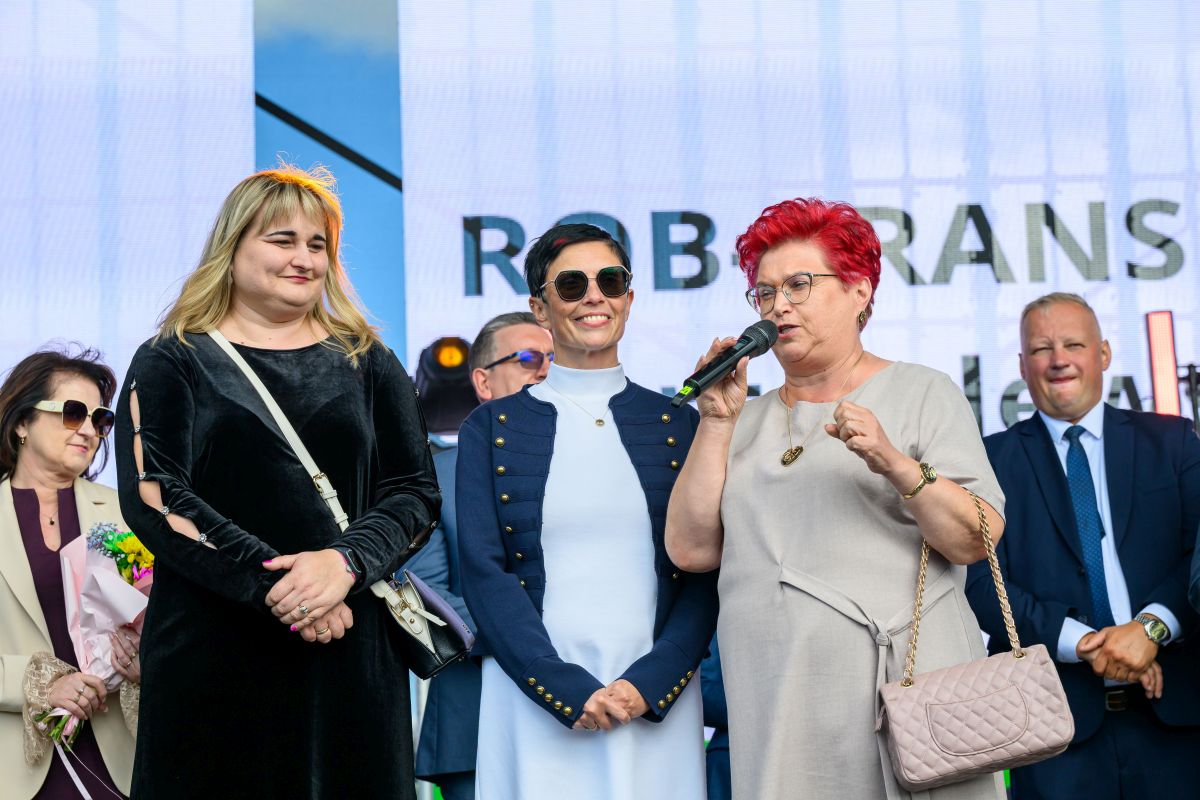 Grupa osób na scenie, jedna kobieta mówi do mikrofonu. Zróżnicowane kolory ubrań, tło z napisem. Atmosfera formalna.