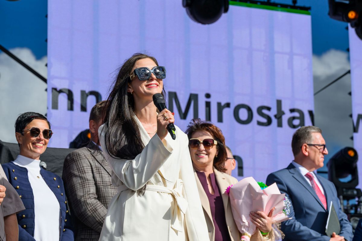 Na scenie stoi kobieta w białym płaszczu z mikrofonem, uśmiecha się. W tle duży ekran z tekstem i niebo. Obok niej kilka osób w formalnych strojach.