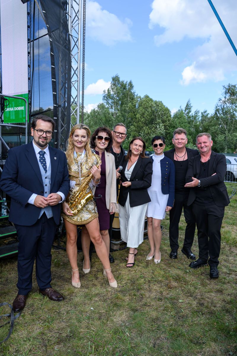 Grupa ludzi na trawie w eleganckich strojach, kobieta trzyma saksofon. W tle sceniczne rusztowanie i drzewa, niebo z chmurami.