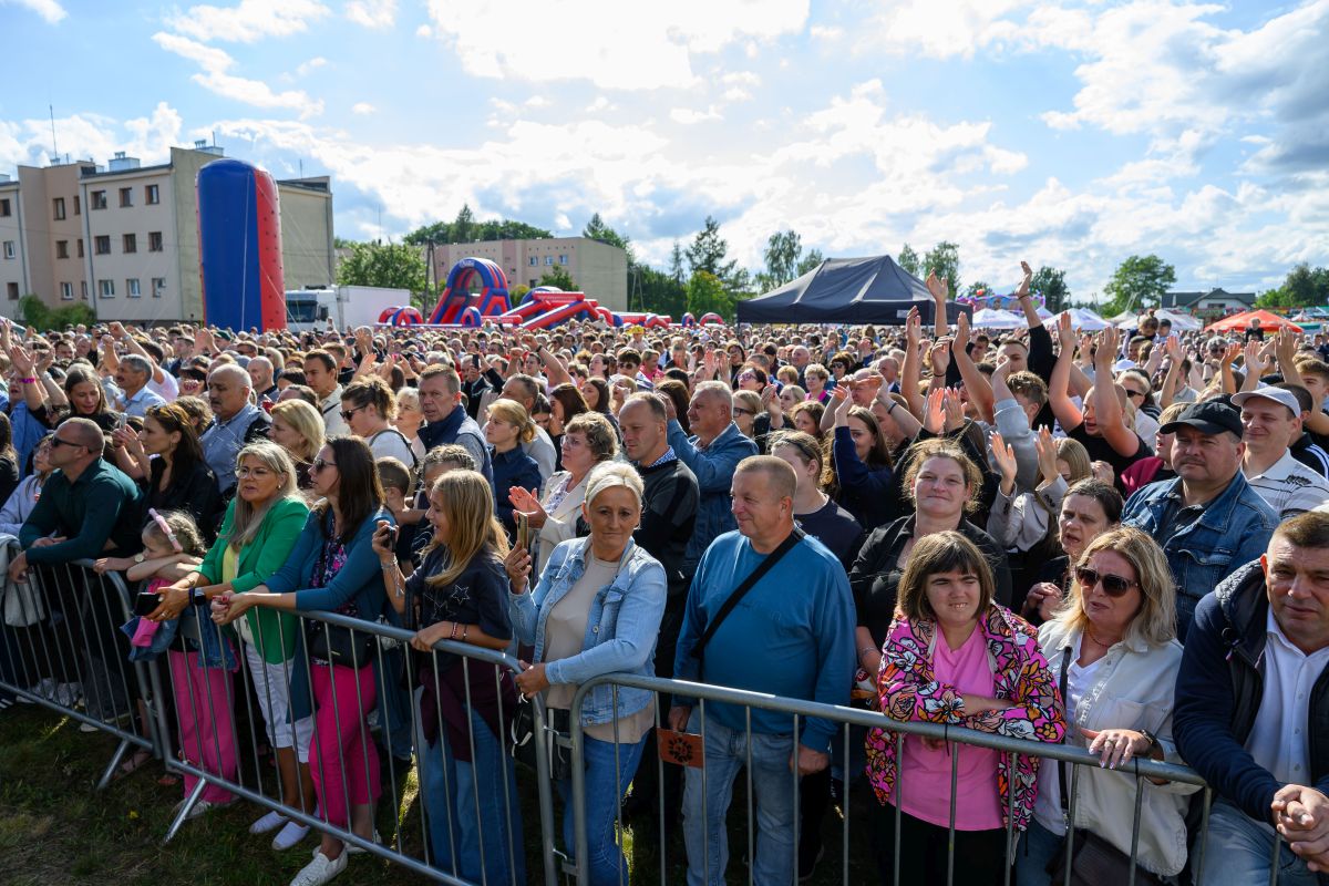 Duży tłum ludzi za barierką na plenerowym wydarzeniu. Jasne niebo z białymi chmurami w tle. Kolorowe dmuchańce i namioty wokół.