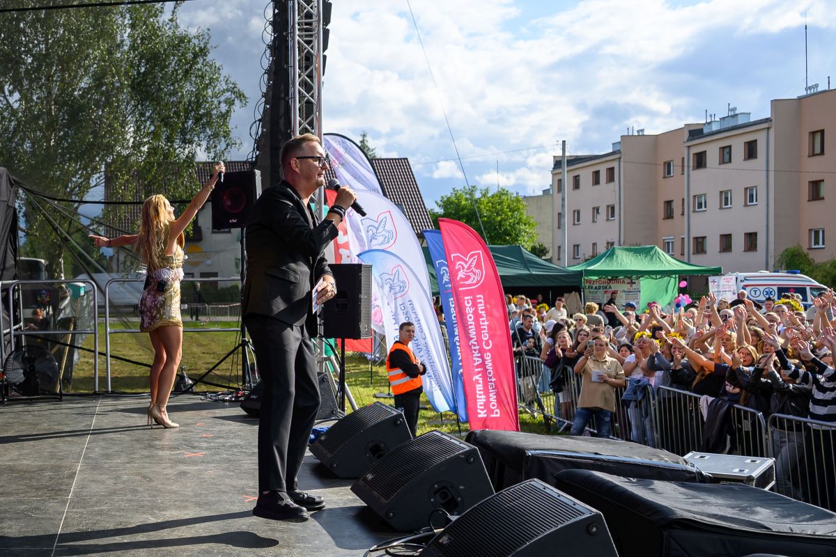 Koncert na scenie plenerowej, mężczyzna w garniturze z mikrofonem, kobieta w sukience, tłum ludzi i budynki w tle.