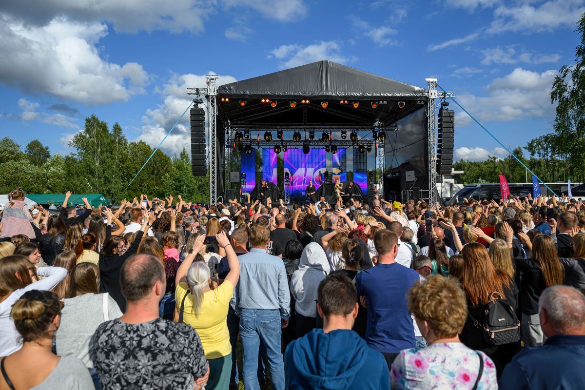 Tłum fanów przed sceną na koncercie plenerowym. Nad sceną czarne zadaszenie, tło z niebieskim niebem i chmurami. Ludzie unoszą ręce, scena oświetlona.