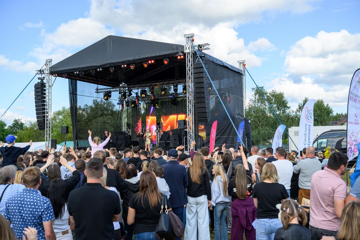 Koncert plenerowy z czarną sceną, kolorowymi światłami i tłumem widzów na tle błękitnego nieba oraz zieleni drzew.