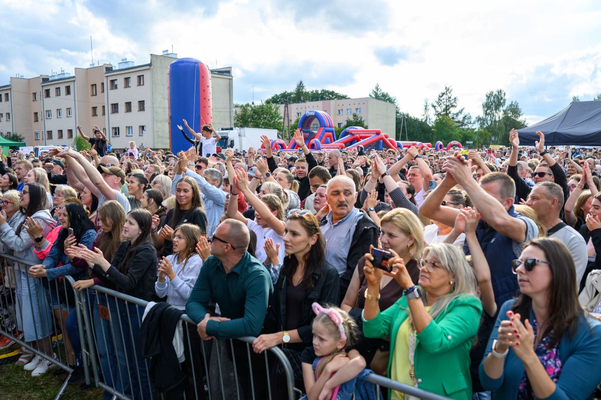 Tłum ludzi na plenerowym koncercie przy jasnym niebie. Kolorowe dmuchańce w tle przy blokach mieszkalnych. Ludzie entuzjastycznie klaszczą.