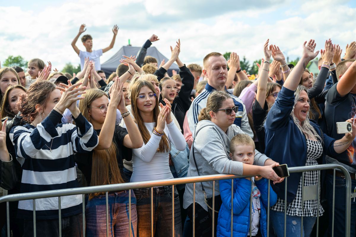 Grupa ludzi na koncercie, wznoszących ręce w radosnym geście. W tle widać niebo pełne chmur. Barwne ubrania tworzą żywą atmosferę.