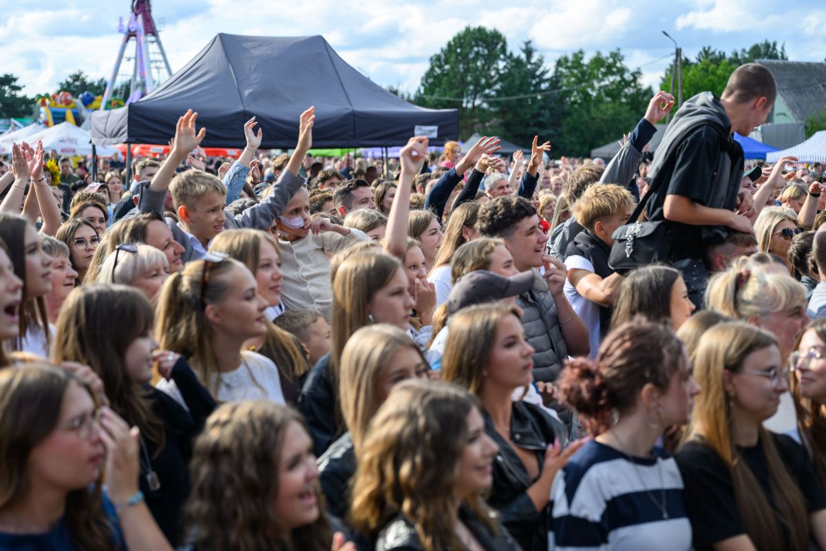 Tłum ludzi na plenerowym wydarzeniu, radosne twarze. Namiot w tle, zielone drzewa. Słoneczny dzień, atmosfera festiwalu.