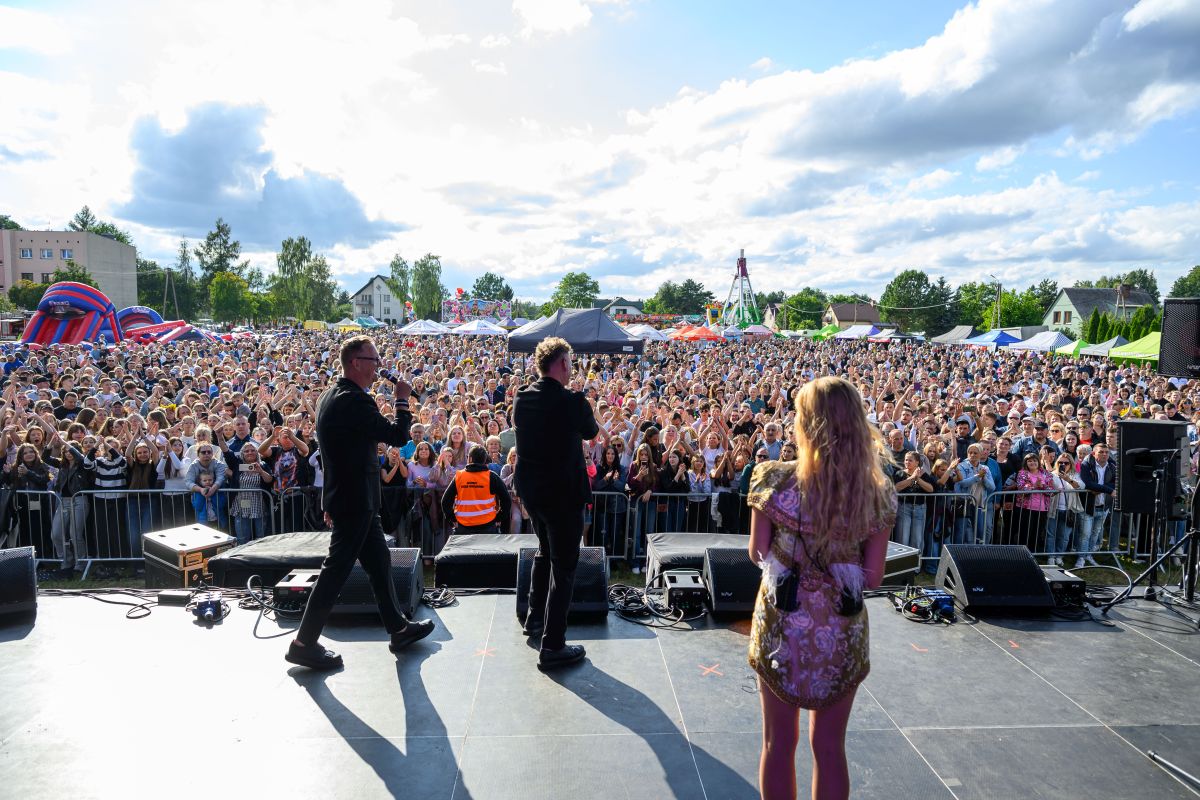 Troje wykonawców na scenie przed tłumem ludzi na festiwalu. Tło wypełnione kolorowymi namiotami, niebo z chmurami. Dominuje zielona sceneria.