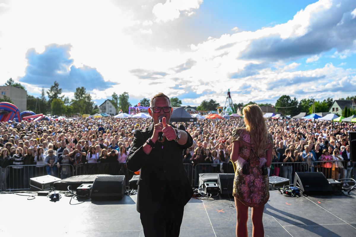 Koncert na świeżym powietrzu; tłum ludzi przed sceną. Słońce przebija się przez chmury na niebieskim niebie. Artysta z mikrofonem i kobieta na scenie.