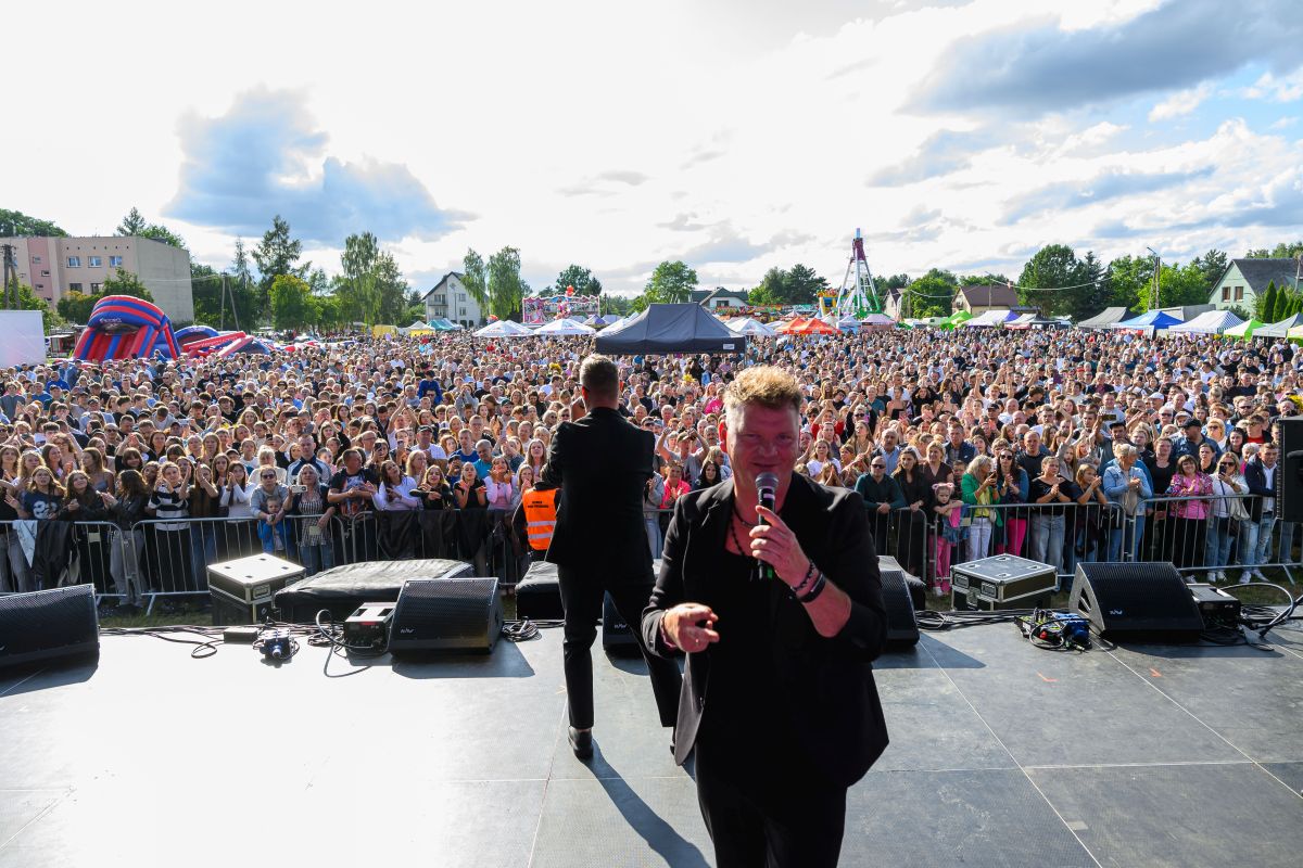 Koncert na świeżym powietrzu z dużą publicznością na tle jasno-niebieskiego nieba. Dwóch artystów w ciemnych ubraniach na scenie. W tle kolorowe namioty i atrakcje.