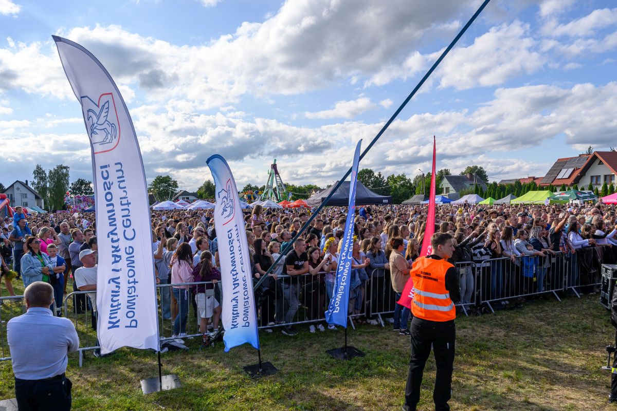 Tłum ludzi na koncercie plenerowym odgrodzonym barierkami. Niebieskie i białe flagi, słoneczny dzień z chmurami. Teren zielony, w tle namioty i domy.