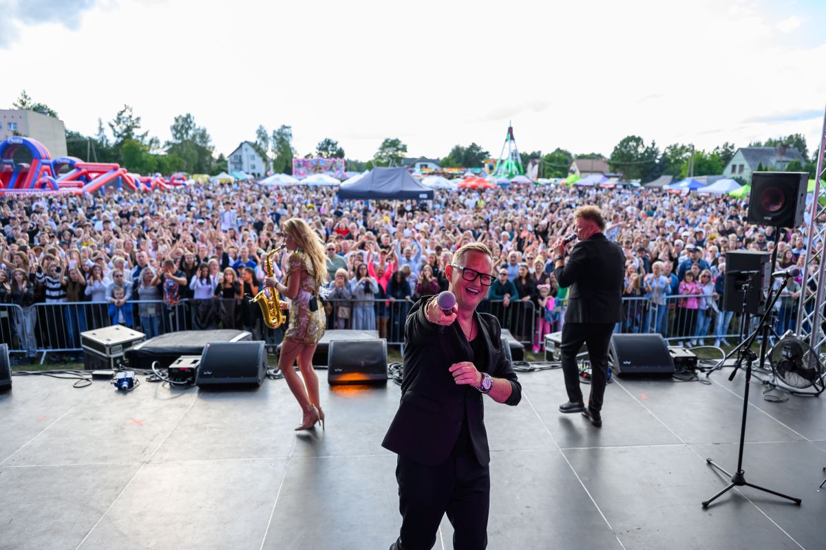Na zdjęciu widzimy koncert na świeżym powietrzu. Muzyk z mikrofonem na scenie w czarnym ubraniu. W tle tłum ludzi i dmuchane atrakcje.