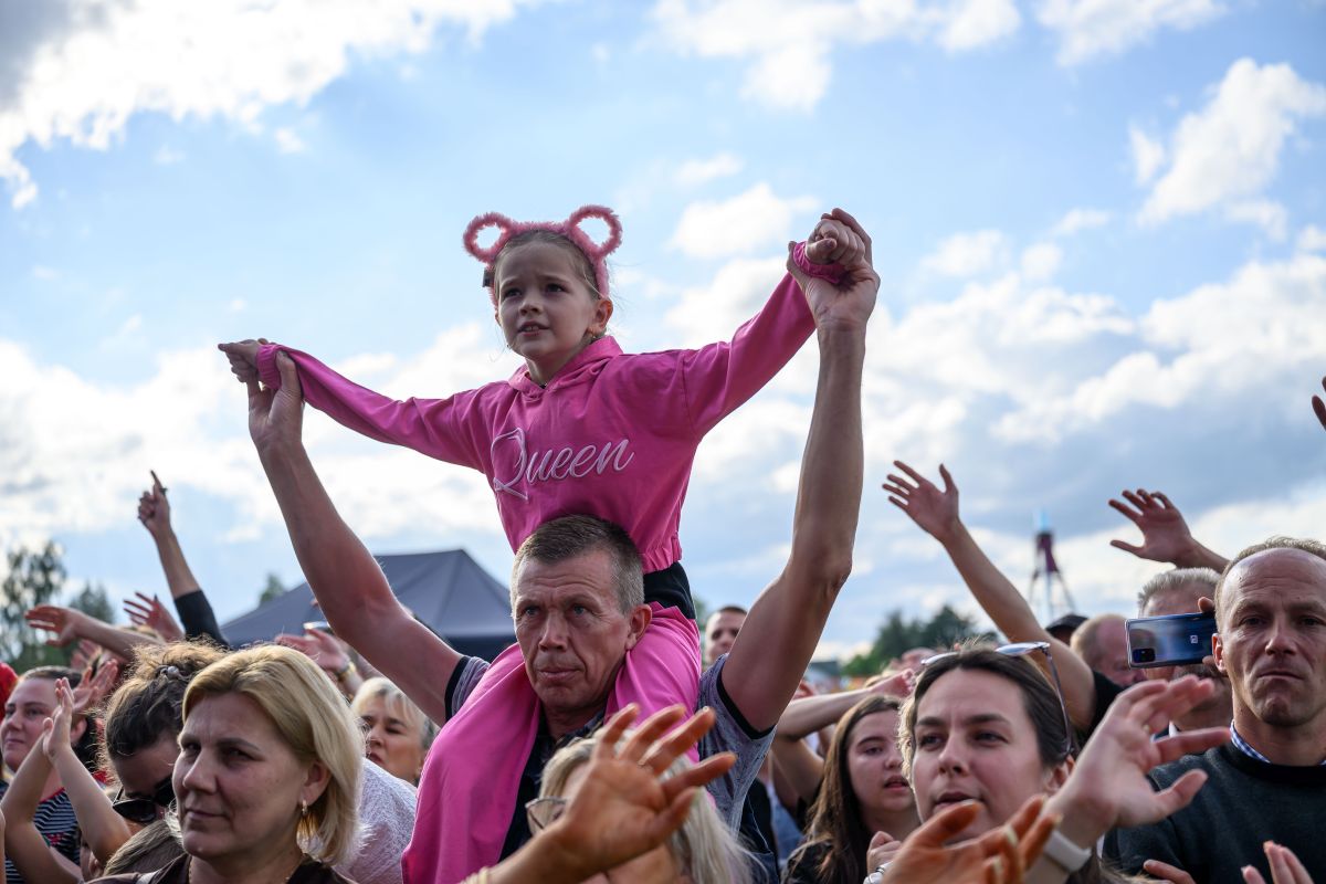 Dziewczynka w różowym ubraniu siedzi na ramionach mężczyzny w tłumie. Tło to błękitne niebo z chmurami. Wokół widać innych uczestników wydarzenia.